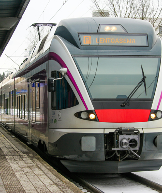 Juna vantaalla pysähtyneenä asemalle. Juna on matkalla lentoasemalle ja taustalla näkyy rakennuksia.